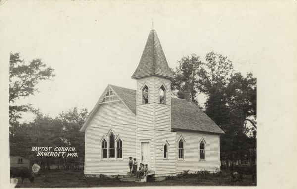 Photographic postcard view of the exterior of the Baptist Church with a bell tower above the entrance. There is a group of children gathered on the front stoop, and a cow grazing in the grass on the left. Caption reads: "Baptist Church, Bancroft, Wis."
