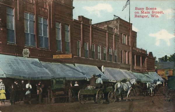 Colorized view across street towards pedestrians and horse-drawn vehicles in front of a row of shops. Caption reads: "Busy Scene on Main Street, Bangor, Wis."