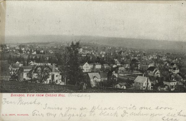 Photographic postcard of an elevated view of Baraboo from a hill, showing mainly dwellings. Bluffs are in the far background. Caption reads: "Baraboo, View from Cheeks Hill, Wis."