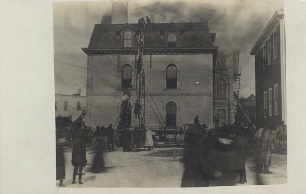 Photographic postcard view of firemen fighting a high school fire. A small crowd of people are watching from the street.
