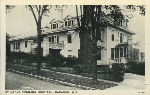 Exterior view of the hospital. A curved driveway leads up to the entrance behind a low stone wall planted with shrubs. Trees line the street. Caption reads: "St. Marys Ringling Hospital, Baraboo, Wis."
