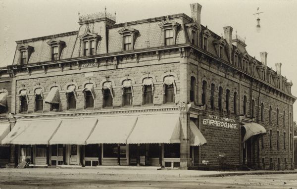 Exterior view from street of the stone hotel on a corner. Large awnings are over the first floor windows. Decorative ironwork is on the roof. Caption reads: "Hotel Warren, Baraboo, Wis."