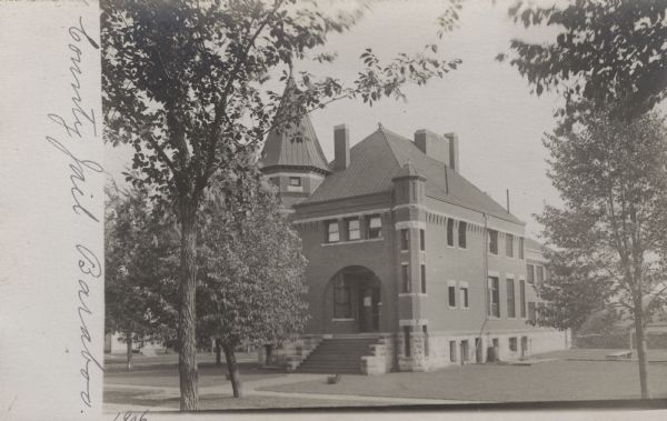 Photographic postcard view of the exterior of the County Jail surrounded by a lawn. A sidewalk leads to wide stone steps that rise up to an arched entrance. There is a turret on the left front corner of the building, and barred windows in the back. Trees line the street.