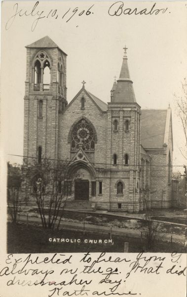 Photographic postcard of an elevated view across street of the front of the Catholic Church, which is a large, stone building. There is a belfry on the left, a steeple on the right, and a Gothic arch and a rose window over the entrance. Caption reads: "Catholic Church."