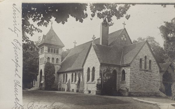 Photographic postcard view of the exterior of the stone church. There are four crosses, one on the bell tower, and three others at the corners of the roof. Windows and entrances are arched, and vines grow along some of the sides of the stone walls.