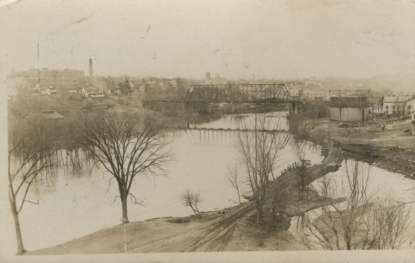 Elevated view of the Baraboo River. The main part of town is on the far left shoreline. On the right industrial buildings are near a bridge spanning the river. In the foreground is a dirt road crossing over a fork in the river.