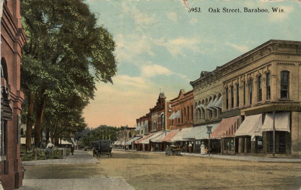 Colorized postcard view down a street in a central business district. There is a park on the left, and storefronts with awnings on the right. A bank is on the corner. Automobiles are parked on the left near a park. Caption reads: "Oak Street, Baraboo, Wis."