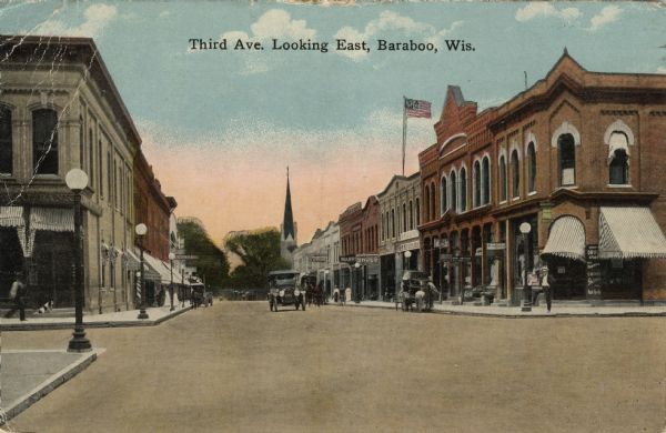 Colorized postcard view of the central business district. The street is lined with businesses and street lamps. A man and a dog are in front of the bank on the left. A car is driving down the street, and horses and carts are parked at the curbs. Caption reads: "Third Ave. Looking East, Baraboo, Wis."