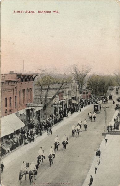Colorized postcard of an elevated view of a parade going through the central business district. People in the parade are wearing helmets and riding horseback behind three carriages. Crowds are gathered on the sidewalks. Caption reads: "Street Scene, Baraboo, Wis."