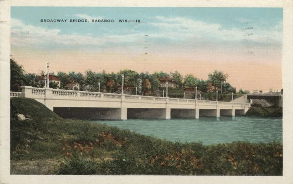 Colorized postcard view from shoreline of a stone bridge lined with lampposts. Dwellings and commercial buildings are on the far side. An elevated railroad bridge crosses the road along the far shoreline. Caption reads: "Broadway Bridge, Baraboo, Wis."