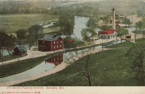 Colorized postcard with an elevated view of the pumping station. On the left is a bridge over the river, and buildings on a strip of land between the river and a stream. Caption reads: "City Water Pumping Station, Baraboo, Wis."