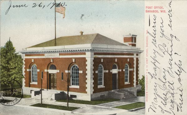 Colorized postcard of a slightly elevated view of the post office. There are wide front steps up to the arched entrance from the sidewalk. The arched entrance is flanked by arched windows. There is a flag on a flagpole on the roof above the entrance. Caption reads: "Post Office, Baraboo, Wis."