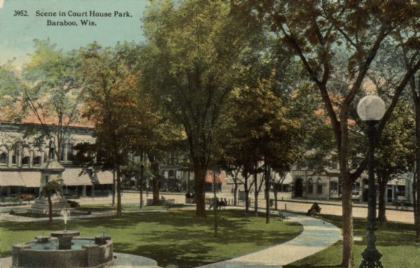 Colorized postcard view of a park in a central business district. There is a fountain, a statue, and benches along the walkway. Caption reads: "Scene in Court House Park, Baraboo, Wis."