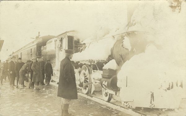 Photographic postcard view of people standing on a platform next to a snow-covered train arriving at a station.