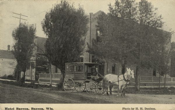 View from street toward a hotel, mostly obscured by trees. A horse and carriage with its driver and passengers is parked at the curb. Caption reads: "Hotel Barron, Barron, Wis."