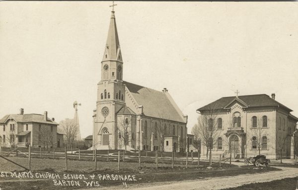 Photographic postcard view of the church, with a parsonage on the left and a school on the right. A fenced-in field is across the street and an automobile is parked at the corner. Caption reads: "St. Marys Church, School & Parsonage, Barton, Wis."