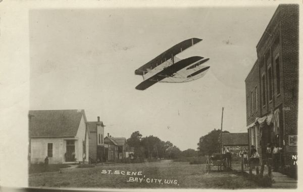 Photographic postcard view of a street in a rural community's central business district. A biplane, printed into the background, is flying low overhead. A group of people are gathered in front of a storefront near a horse and buggy parked at the curb. Caption reads: "Street Scene, Bay City, Wis."