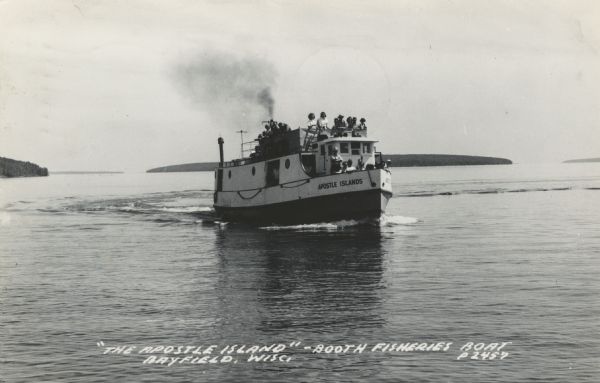 Photographic postcard view of a fishing boat in Lake Superior with  tourists on board. There is an island on the horizon. Caption reads: "'The Apostle Island' Booth Fisheries Boat, Bayfield, Wisc."