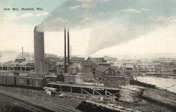 Elevated view of the saw mill. Lumber is stacked in front of several buildings and railroad tracks. Caption reads: "Saw Mill, Bayfield, Wis."