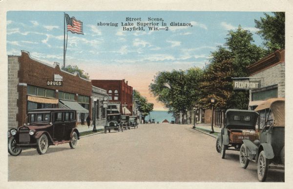 Colorized postcard view down a street (Rittenhouse Avenue) to Lake Superior. The street is lined with businesses and parked cars. Caption reads: "Street Scene, showing Lake Superior in distance, Bayfield, Wis."
