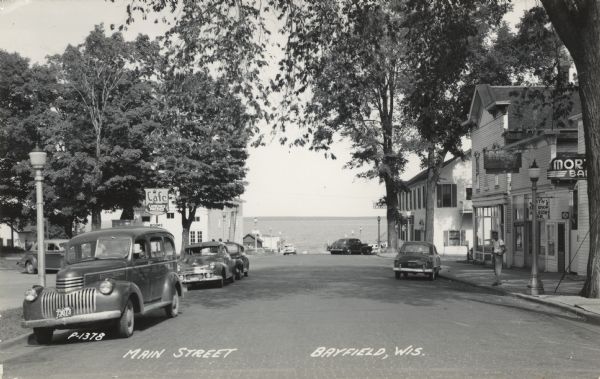Photographic postcard view down Main Street, with Lake Superior in the distance. There is a cafe on the left and bars on the right. Cars are parked along the curbs. Caption reads: "Main Street, Bayfield, Wis."