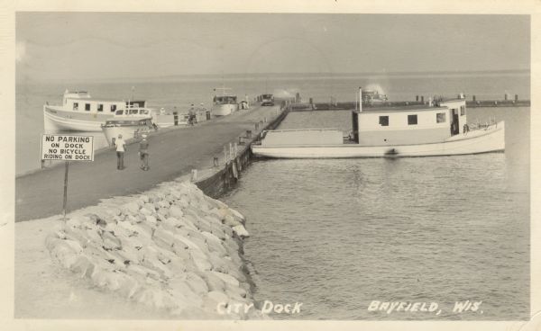Photographic postcard view of the City Dock on Lake Superior. There are four boats, and people and cars are on the dock. A sign in the foreground reads: "No Parking on Dock, No Bicycle Riding on Dock." Caption reads: "City Dock, Bayfield, Wis."
