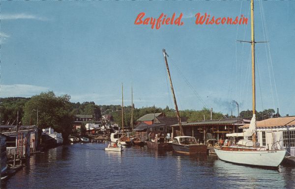 Photographic postcard view from water of a marina. Sailboats and motorboats are docked. More boats are stored up on the shoreline. Trees and buildings are in the background. Caption reads: "Bayfield, Wisconsin."