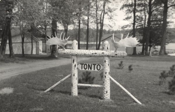 Photographic postcard view of cabins by a lake. In the foreground is a birch sign with antlers that reads: "Tonto."