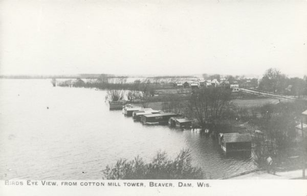 Photographic postcard of an elevated view of a lake with boathouses along the shore and dwellings in the distance. Caption reads: "Birds Eye View from Cotton Mill Tower, Beaver Dam, Wis."