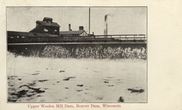 Postcard view of a dam with a mill behind it. Caption reads: "Upper Woolen Mill Dam, Beaver Dam, Wisconsin."
