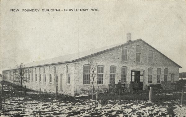 Photographic postcard view  of a foundry. There are workmen standing near the entrance. Caption reads: "New Foundry Building - Beaver Dam - Wis."