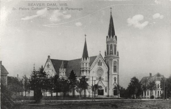 Photographic postcard view of the Catholic Church and Parsonage. There is a rose window over the entrance of the cathedral. Crosses are on the church towers. Caption reads: "Beaver Dam, Wis. Peters Catholic Church & Parsonage."