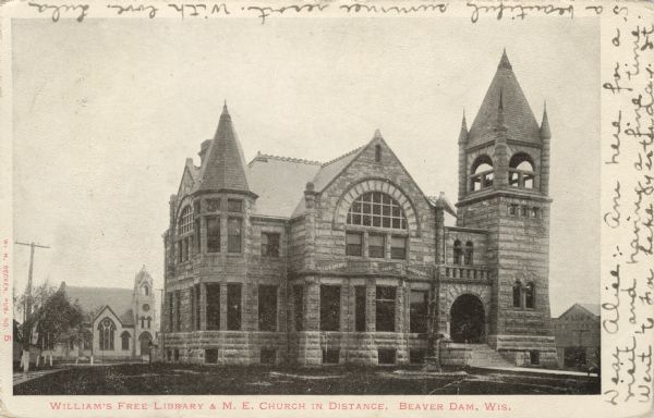 Photographic postcard of an exterior view of the library. The library is a stone building with an arched entrance and arched windows on the second floor. The Methodist Episcopal Church is on the block behind the library. Caption reads: "Williams Free Library & M.E. Church in Distance, Beaver Dam, Wis."