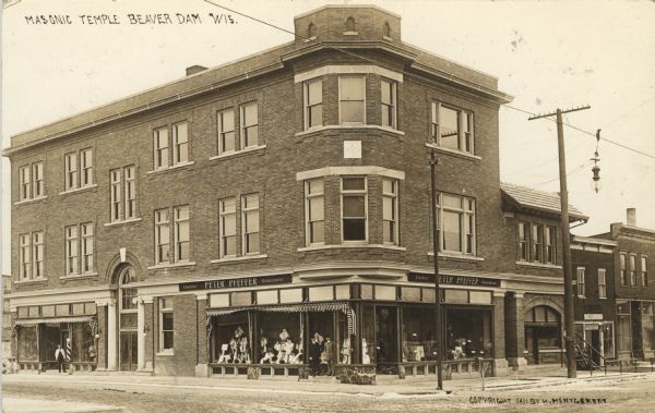 Photographic postcard of an exterior view from the street of the Masonic Temple on a corner. The Peter Pfeffer haberdasher shop is on the ground floor at the corner, and two men stand at the entrance. On the left on the other side of the temple entrance, a man stands in front of a barbershop. To the right of the temple is a building with a bicycle repair shop. Caption reads: "Masonic Temple, Beaver Dam, Wis."