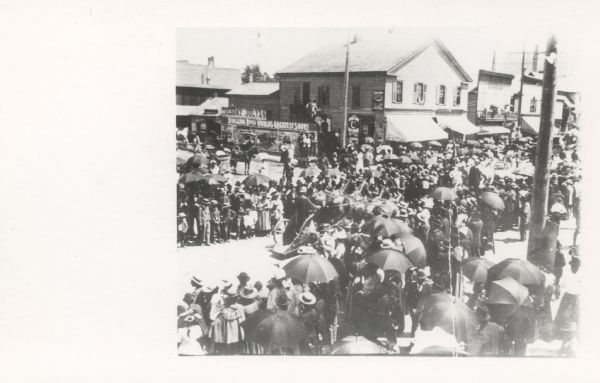Photographic postcard of an elevated view of the Circus Parade on a rainy day. Crowds of people line the street, and many are holding umbrellas.