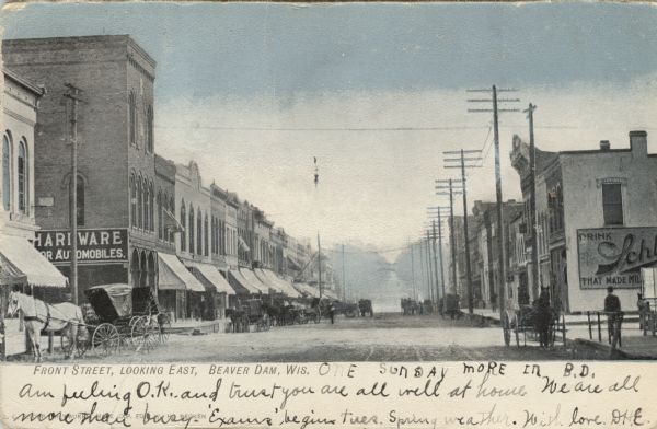 Colorized postcard view down center of the street in the central business district. Horses and buggies are parked along the curbs, and pedestrians are on the sidewalk.