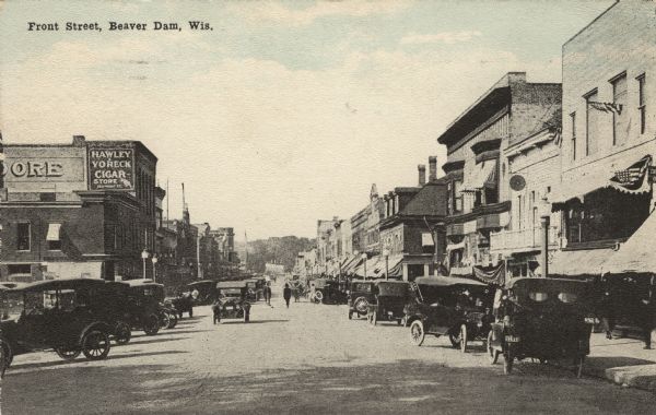 Colorized postcard view down the center of a busy street in the central business district. Many cars are parked at an angle along the curbs. Lampposts line the street. Caption reads: "Front Street, Beaver Dam, Wis."