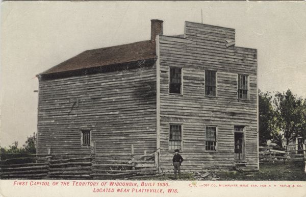 Colorized photographic postcard view of the First Capitol, which is an unpainted wood frame building, with a log fence and a man standing in front. Caption reads: "First Capitol of the Territory of Wisconsin, Built 1836, Located near Platteville, Wis."