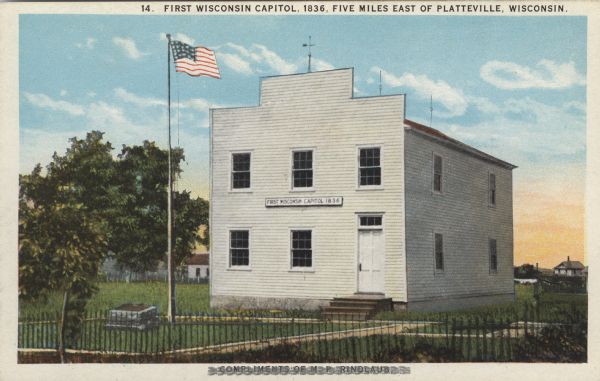 Colorized view of the First State Capitol, which is a wood frame building, painted white, with a flag and a plaque in front. Caption reads: "First Wisconsin Capitol, 1836, Five Miles East of Platteville, Wisconsin."