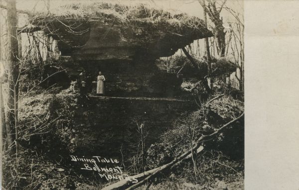Photographic postcard view of a rock formation called "Dining Table" with two people posing next to it. Caption reads: "Dining Table, Belmont Mounds."