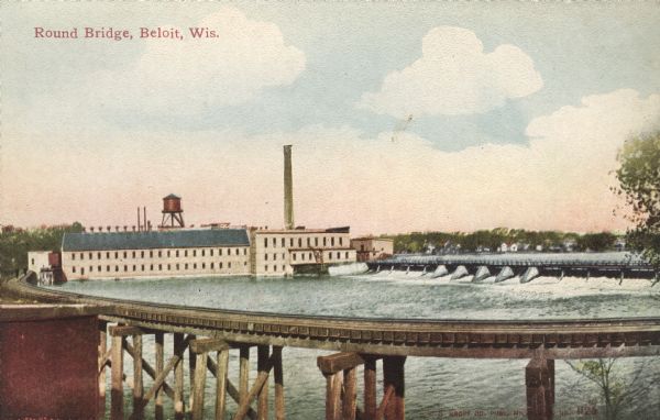 Colorized postcard of an elevated view of the Round Bridge, part of the C.M. and St. Paul R.R. A factory and dam are in the background and dwellings are on the far shore. Caption reads: "Round Bridge, Beloit, Wis."