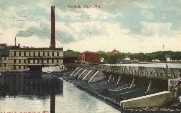 Colorized postcard of an elevated view across a dam towards a factory on the Rock River. Dwellings are on the far bank on the right. Caption reads: "The Dam, Beloit, Wis."