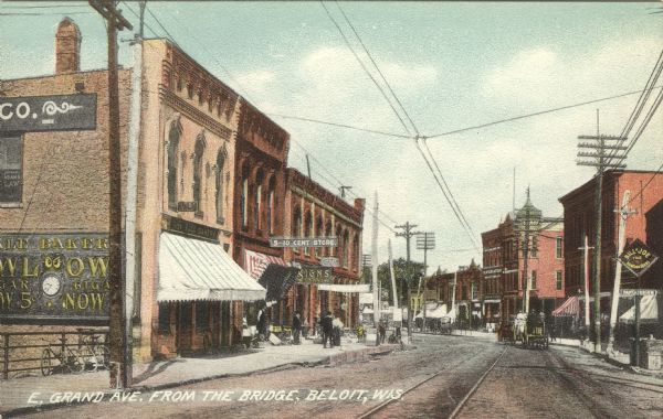 Colorized postcard view looking down street in the central business district. Streetcar tracks run down the center of the street, and horse-drawn vehicles are on the right. Bicycles are parked on a railing on the left. Caption reads: "E. Grand Ave. from the Bridge, Beloit, Wis."