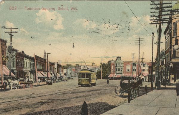 Colorized postcard view of a square in the central business district. A cable car runs down the center of the street, and horse-drawn vehicles are parked along the curbs. Caption reads: "Fountain Square, Beloit, Wis."