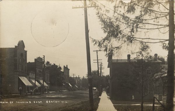 Photographic postcard view of a central business district. On the left is a bank, and a "Place to Eat." Horses and buggies are further down the block.