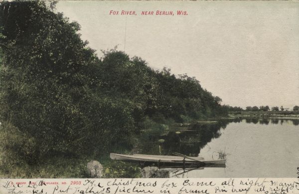 Colorized postcard view along the Fox River. A small rowboat is tied up at the shore. Caption reads: "Fox River, near Berlin, Wis."