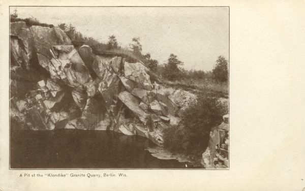 Photographic postcard view of a quarry with water at the bottom. Caption reads: "A Pit at the 'Klondike' Granite Quarry, Berlin, Wis."