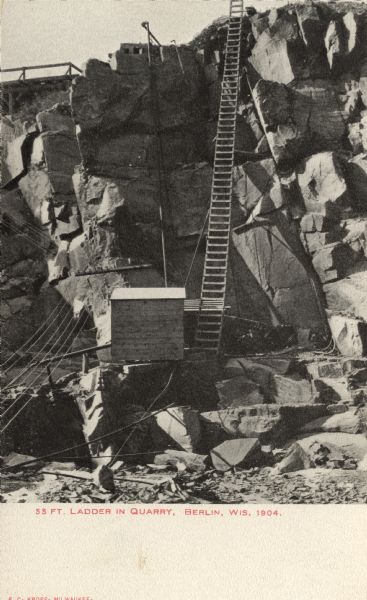 Photographic postcard view of the bottom of the quarry, with a ladder from the bottom of the pit to top. There is a shed at the foot of the ladder. Caption reads: "55 Ft. Ladder in Quarry, Berlin, Wis."