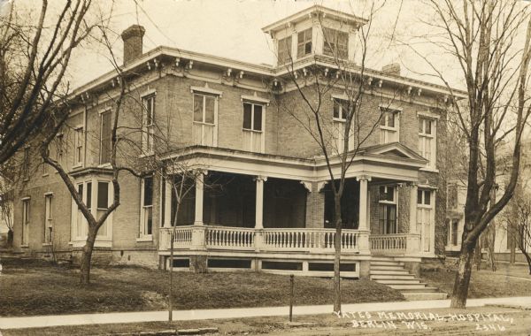 Photographic postcard of an exterior view from the street of a hospital with a large porch. Caption reads: "Yates Memorial Hospital, Berlin, Wis."