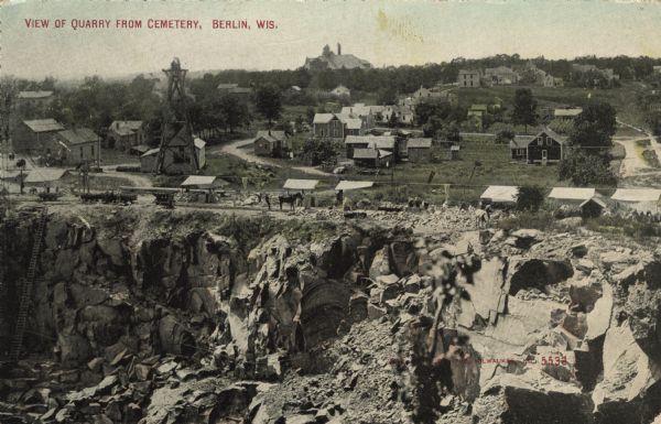 Partially colorized view looking down at a quarry. On the other side of the quarry is the town of Berlin, with dwellings and commercial buildings. Caption reads: "View of Quarry from Cemetery, Berlin, Wis."